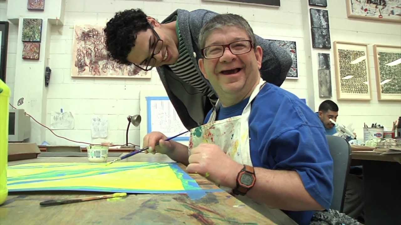 The photo was taken in an art studio setting. A person is sitting at the art table with a yellow piece of paper in front of him as he holds a paint brush and wears a paint splattered apron, orange wrist watch, blue tee shirt, black rimmed glasses as he smiles at the camera. The art teacher is looking over the shoulder of the student wearing a grey and white striped shirt under a grey blazer, with dark rimmed glasses as he smiles at his students work.