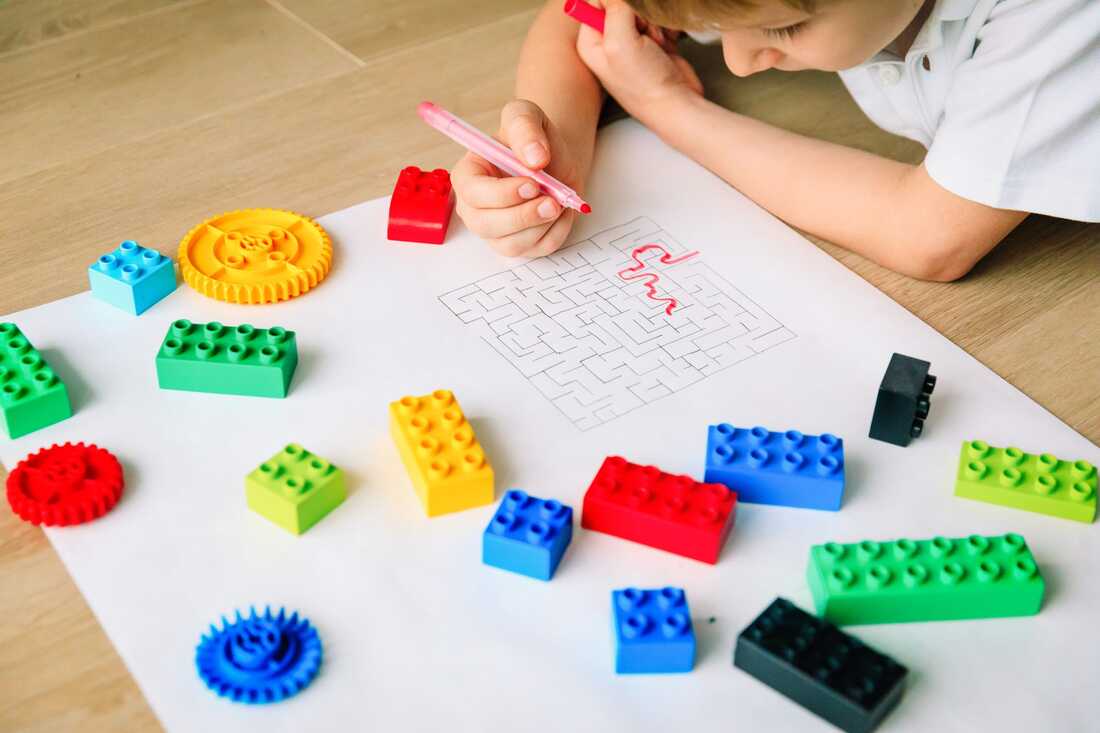 Young boy wearing a white tee shirt laying on a wooden floor, with a piece of paper covered in Lego pieces. The boy is holding a red marker completing a maze puzzle. 