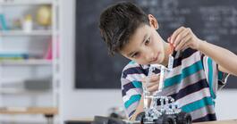 A boy in a black, grey, and blue striped collared shirt sitting at table in a classroom setting. The boy is putting together what looks like a vehicle using Lego type blocks.