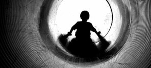 A black and white image of a young child sitting a the end of a tube slide with his back to the camera.