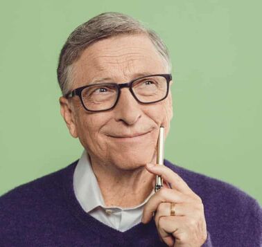 A photo of Bill Gates sitting in front of a green wall. He is wearing black rimmed glasses, a off white collared shirt under a dark blue cardigan with a pen in his hand that bears a gold wedding band. He is looking up to the top right with a smirk on his face holding the bottom of his pen against his cheek.