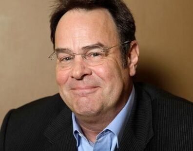 A photo of Dan Aykroyd. He is sitting in front of a tan wall, wearing a light blue button up shirt under a black pinstripe blazer. He is fair skinned, and wearing frameless glasses. 