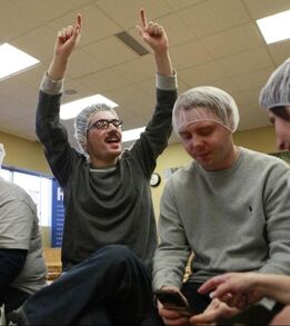 Two boys sitting on a metal table wearing gray long sleeve shirts and hair nets. One of the men is wearing black framed glasses and has his hands in the air and smile.