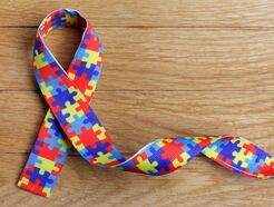 A ribbon with a dark blue, light blue, red, and yellow puzzle piece pattern printed on it. The ribbon is twisted in a support shape and is laying on top of what looks like a wooden table top.