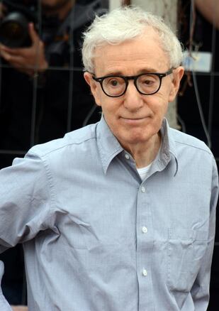 Photo of Woody Allen. Older gentleman with white hair, a light grey button up shirt, and black rimmed sunglasses. 
