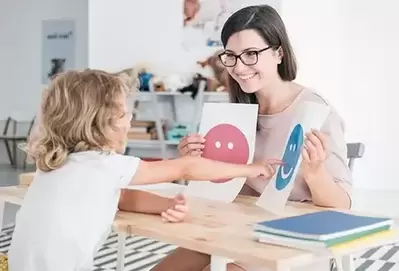 A classroom like setting with a small child with shoulder length curly blond hair sitting across from a young therapist with dark hair and glasses. The therapist is holding two pieces of paper, one with a red frowning emoji and one with a blue smiling emoji. The child is pointing at the blue smiling emoji photo.