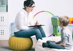 A woman and young boy sitting in a therapeutic setting. The woman is sitting on a yellow knitted floor cushion holding a pencil and a clipboard while she leans forward looking at the child. The child is sitting on a white bean bag chair looking at the therapist.