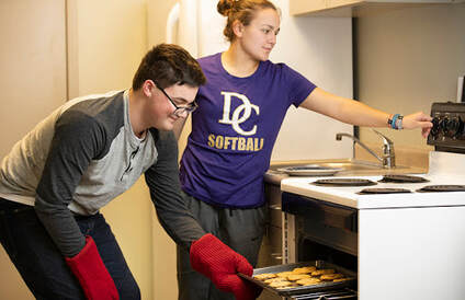Two teenage/young adults standing in a kitchen. The male is wearing jeans, a light and dark grey long sleeve shirt with red oven mitts. He is bent over slightly holding a tray of cookies pulling them out of the oven. The woman in the picture is wearing a dark purple tee shirt with 
